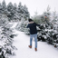 A man carries a freshly wrapped Christmas tree on his shoulder through a snowy tree farm surrounded by snow-covered evergreens.