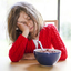 A child in a red jumper leaning their head in their hand and trying to eat a bowl of cereal. 