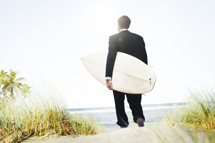 Man in suit with a surfboard on the beach.
