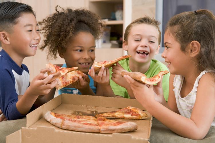 Four young children indoors eating pizza smiling.
