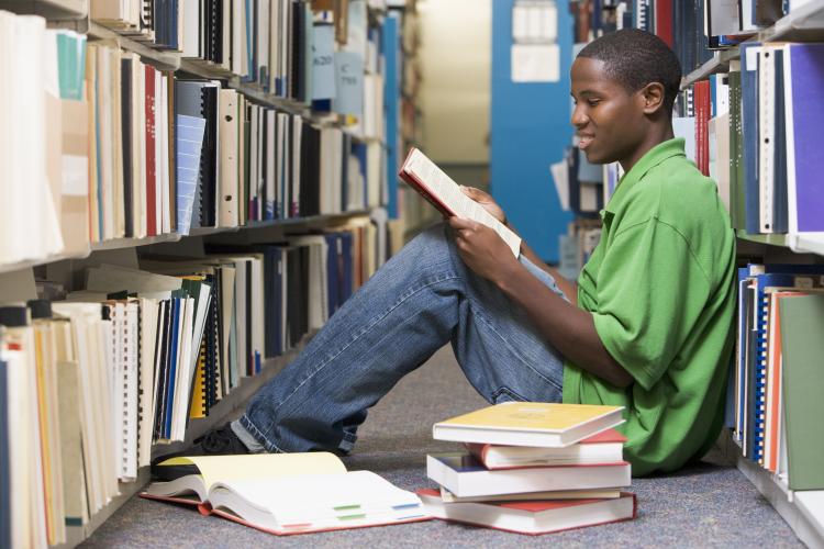 Man sitting on floor in library reading book.