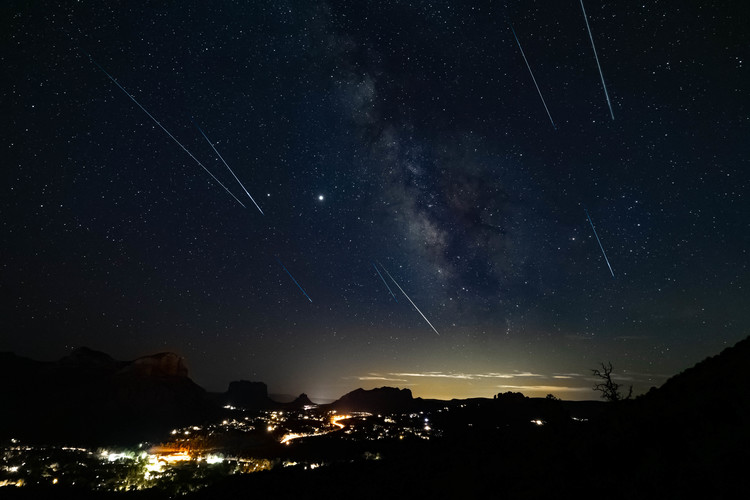 Dark sky with stars and several meteors over a landscape with city lights.