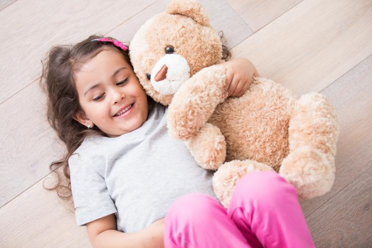 Girl with teddy bear lying on wooden floor at home.