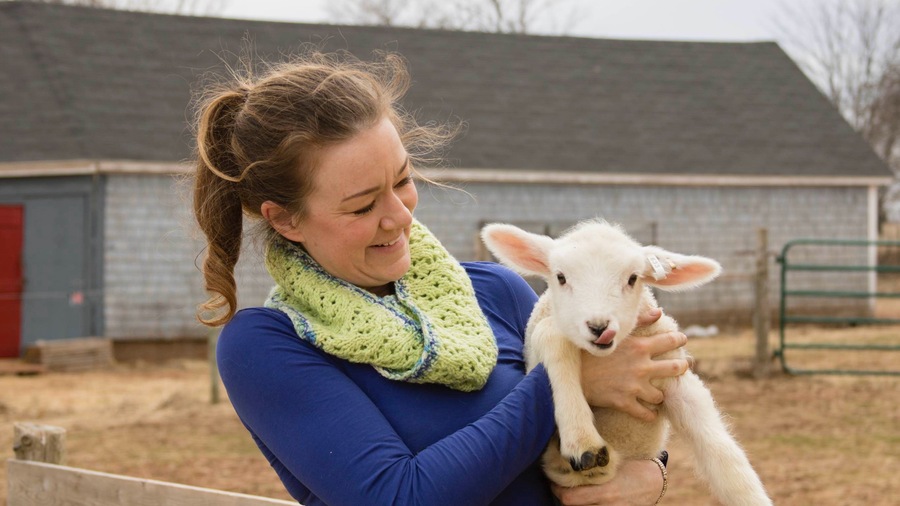 Female farmer holding a lamb licking its mouth. 