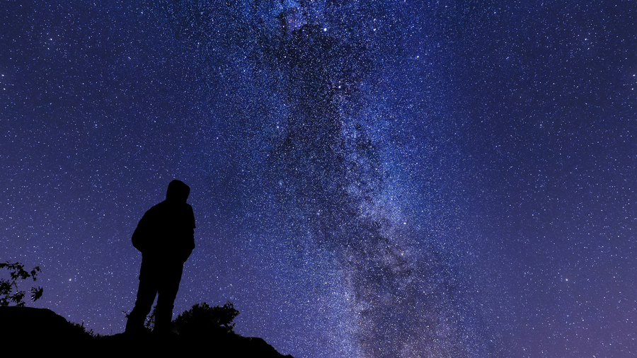 A person standing on a rocky outcrop looking up at the Milky Way in a dark and starry sky.