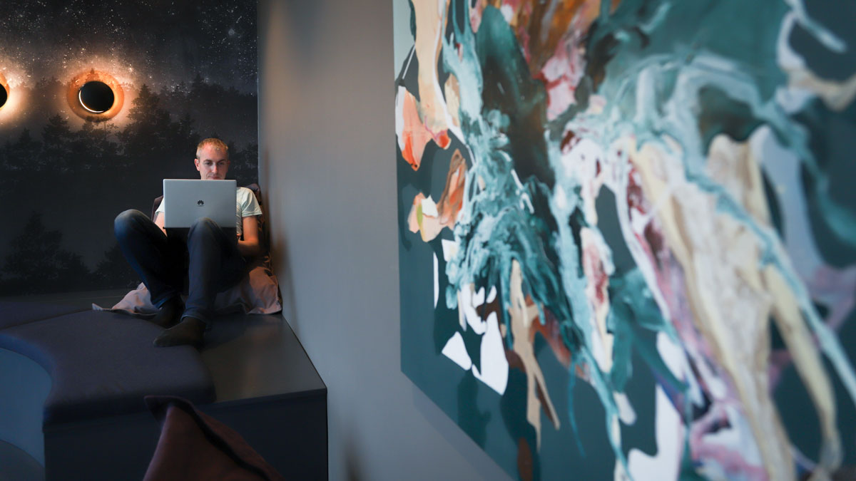 Junger Mann mit weißem T-Shirt arbeitet entspannt auf einem großen Sitzkissen. An der Wand sind abstrakte Malerei zu sehen.